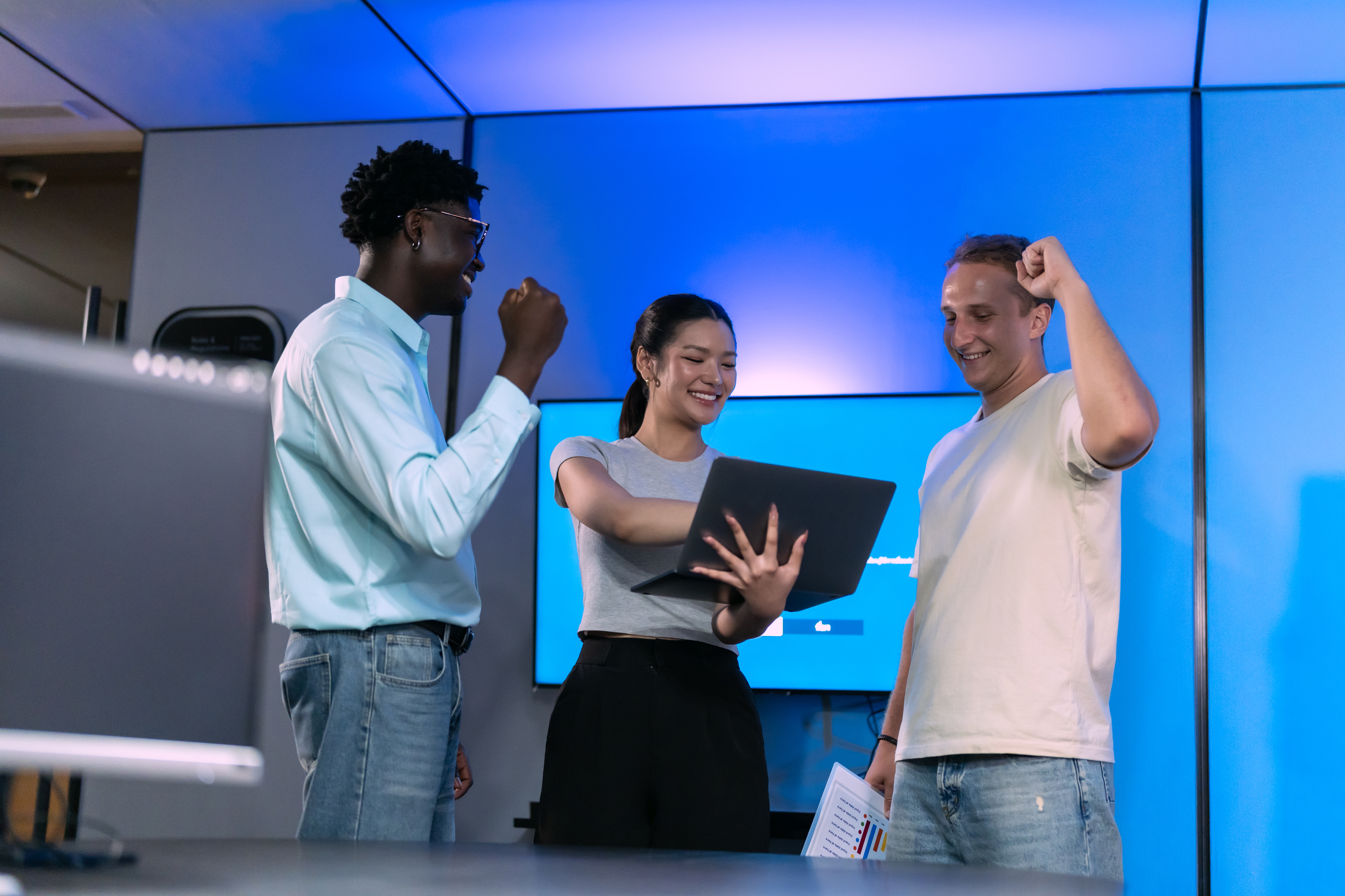 Asian woman holds a laptop while two teammates raise their fists to celebrate a result in a blue lit technology office.
